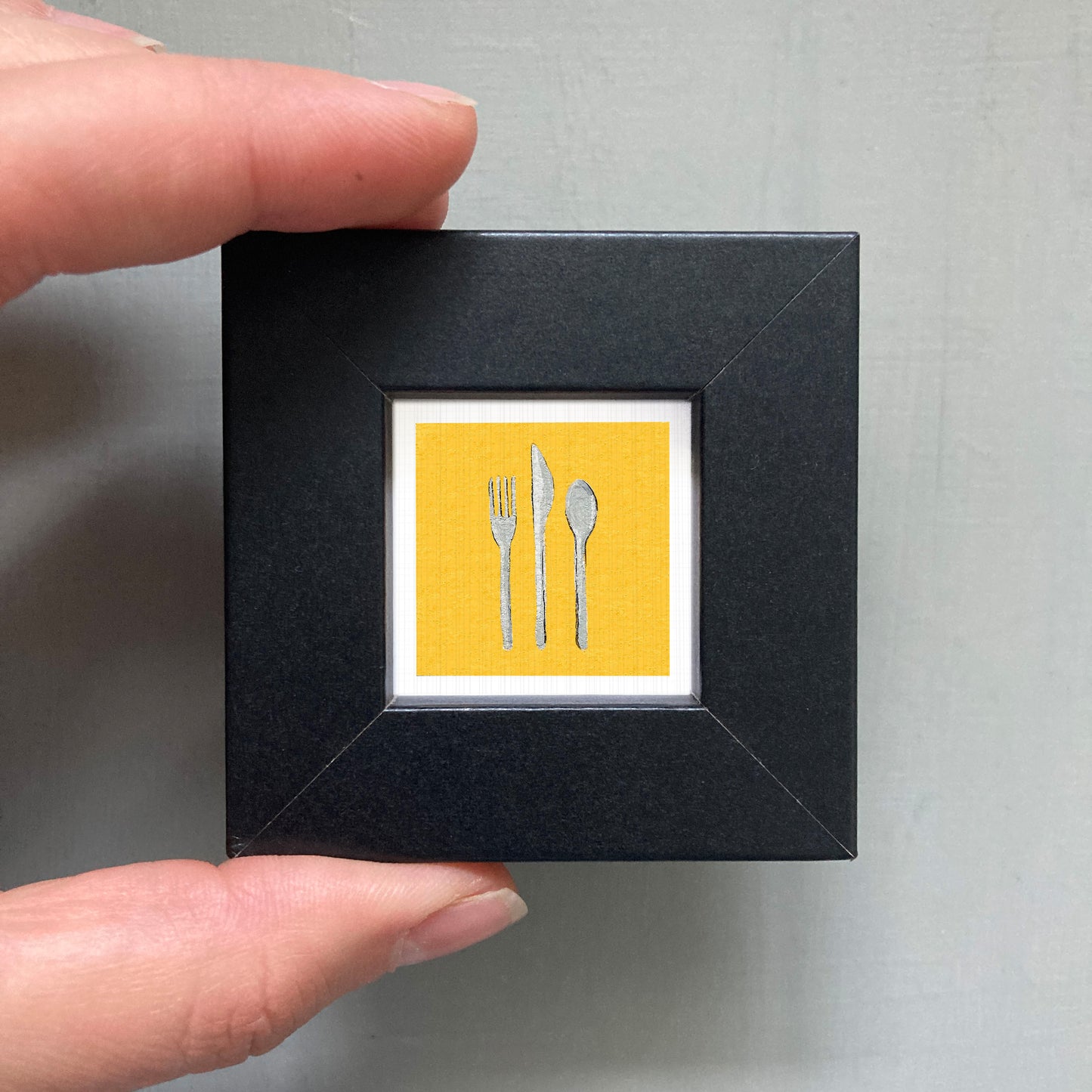 A person's hand is holding a small black frame containing a framed image of a yellow square with a white border, depicting three silverware items - a fork, a knife, and a spoon - against a white background.