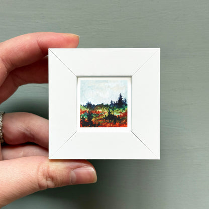 A hand is holding a small white square frame containing a painting of a landscape with trees and a blue sky.