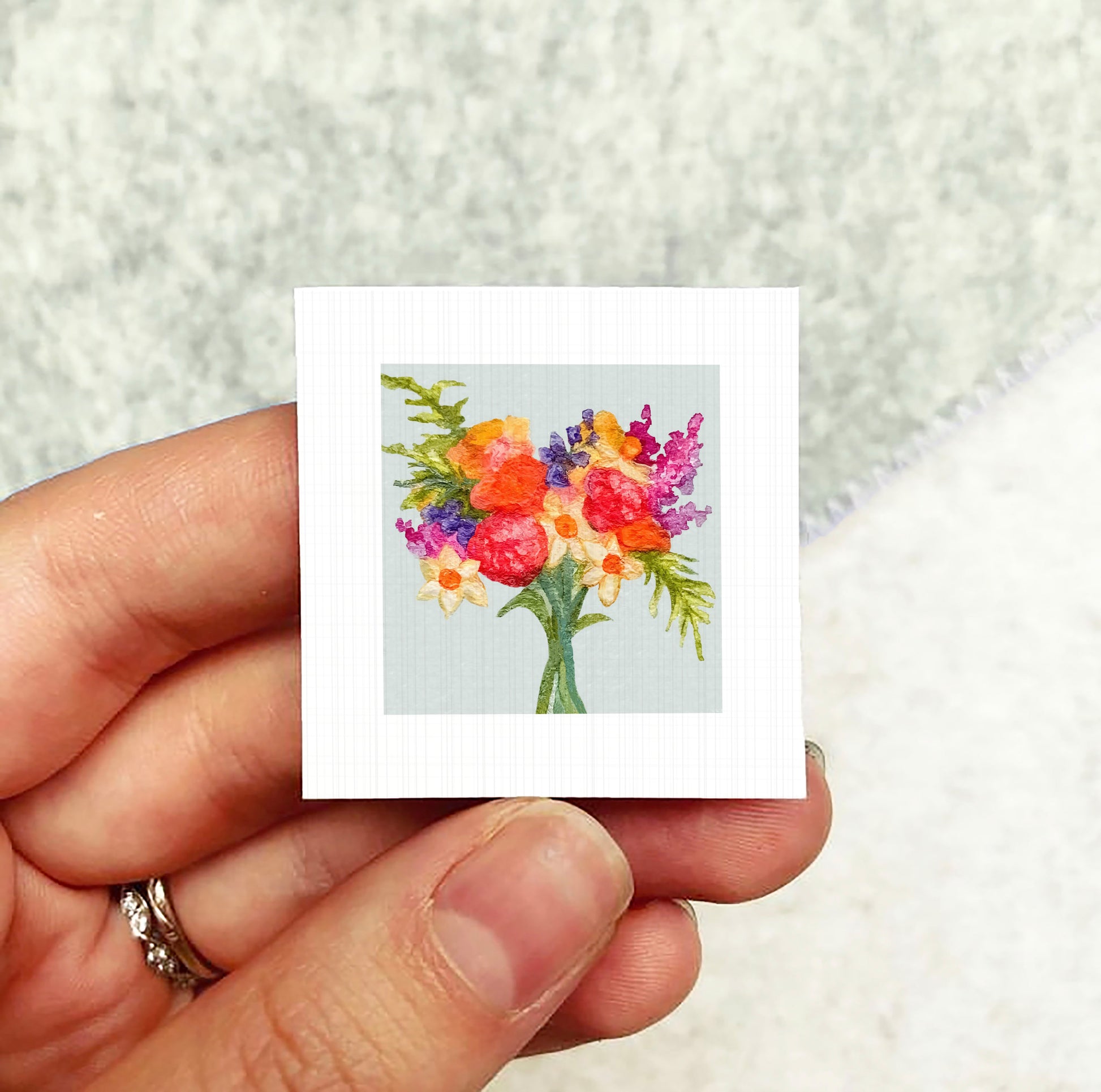 A hand holding a small square card with a colorful floral design on it.