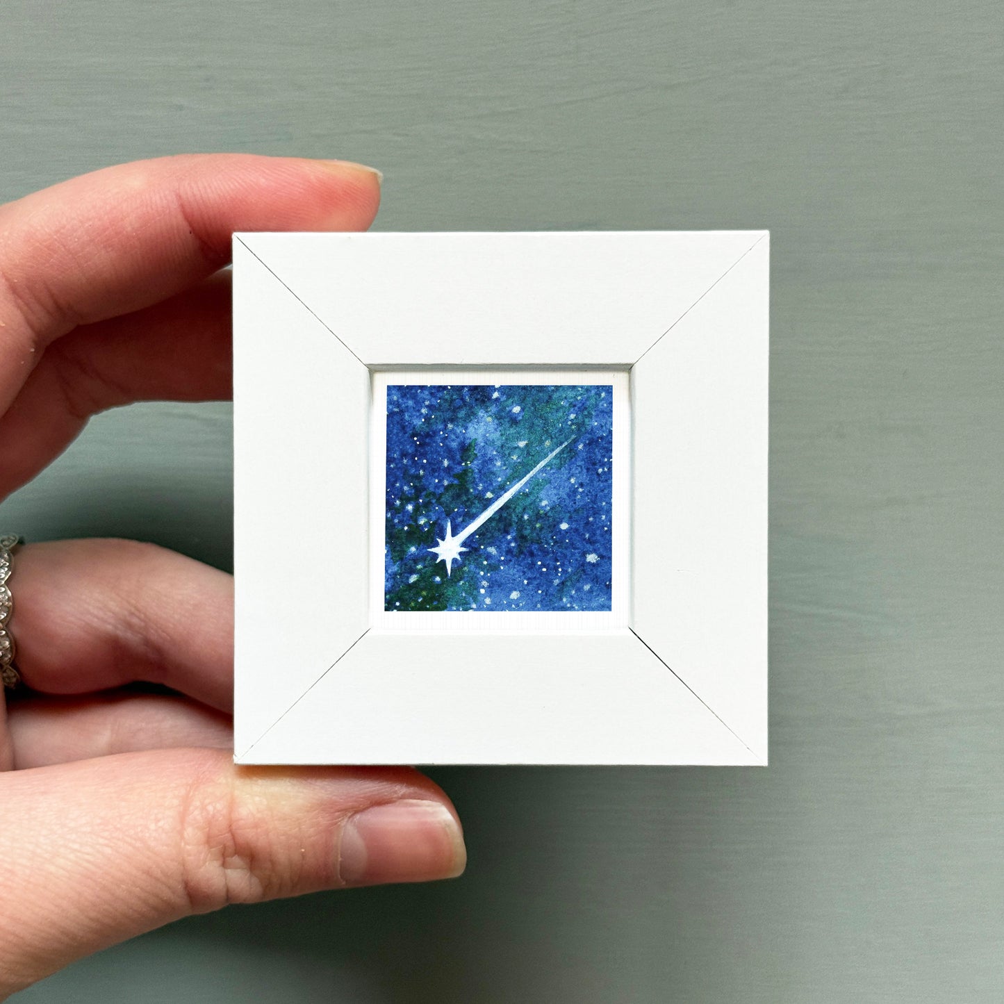 A hand is holding a small white square frame containing a painting of a starry night sky.