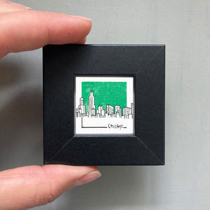 A person's hand is holding a small square frame containing an illustration of a city skyline with a green background.