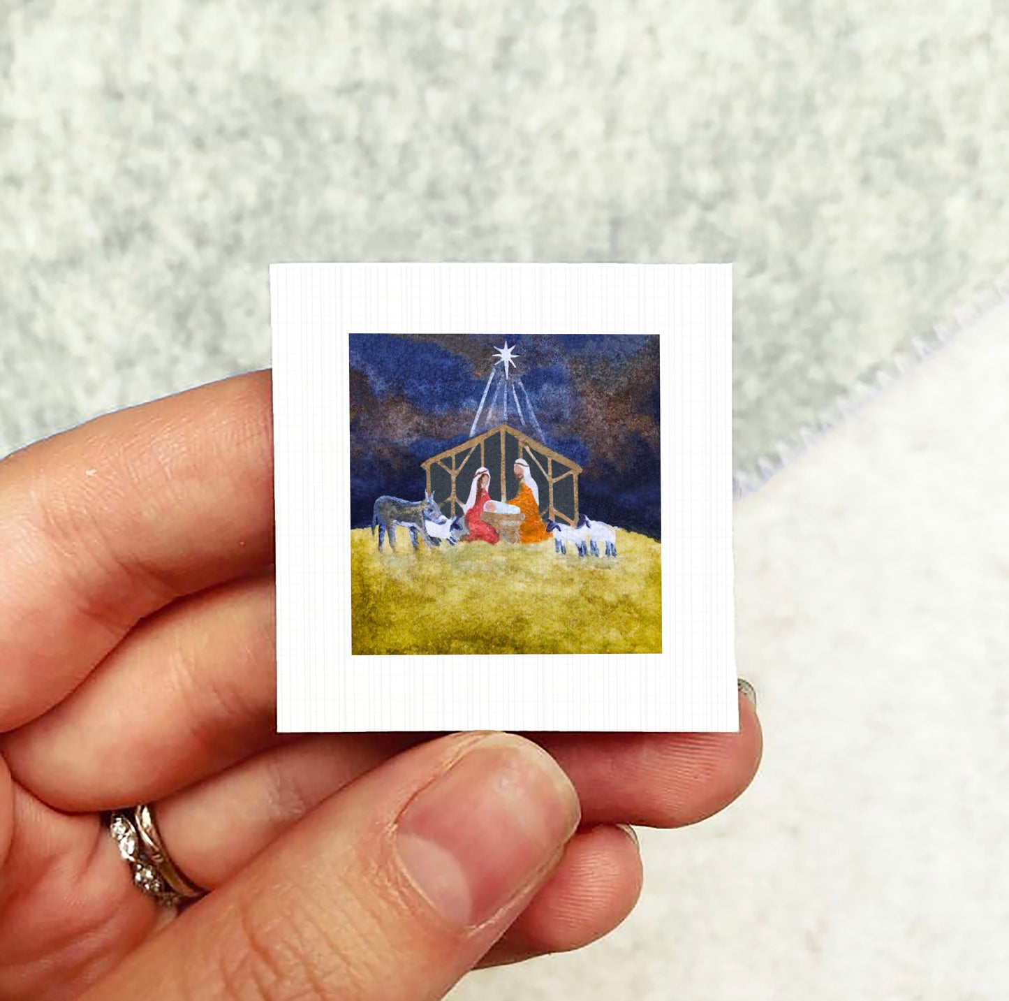A hand holding a small square-shaped picture depicting a nativity scene with a man, a woman, and animals in a stable.