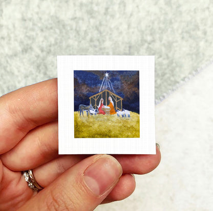 A hand holding a small square-shaped picture depicting a nativity scene with a man, a woman, and animals in a stable.