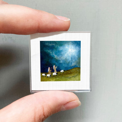 A person's hand is holding a small square picture depicting a pastoral scene with a shepherd and his flock of sheep.
