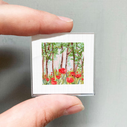A person's hand holding a small square picture of a forest scene with red flowers.