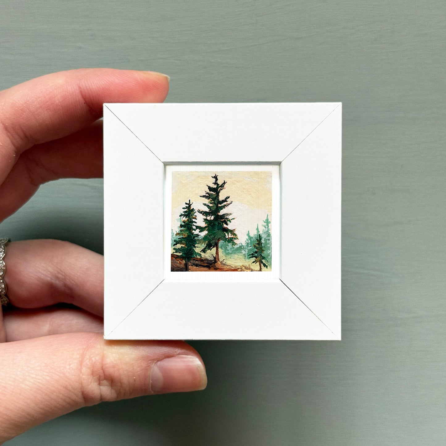 A hand is holding a small white square frame containing a painting of a forest scene with pine trees.