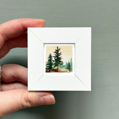 A hand is holding a small white square frame containing a painting of a forest scene with pine trees.