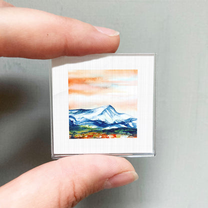 A person's hand is holding a small, square-shaped picture of a mountain landscape with a colorful sky.