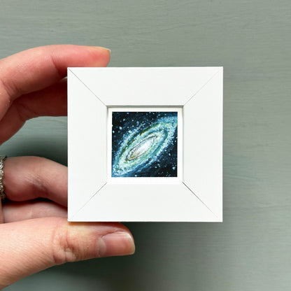 A hand is holding a small white square frame containing a miniature representation of a spiral galaxy.
