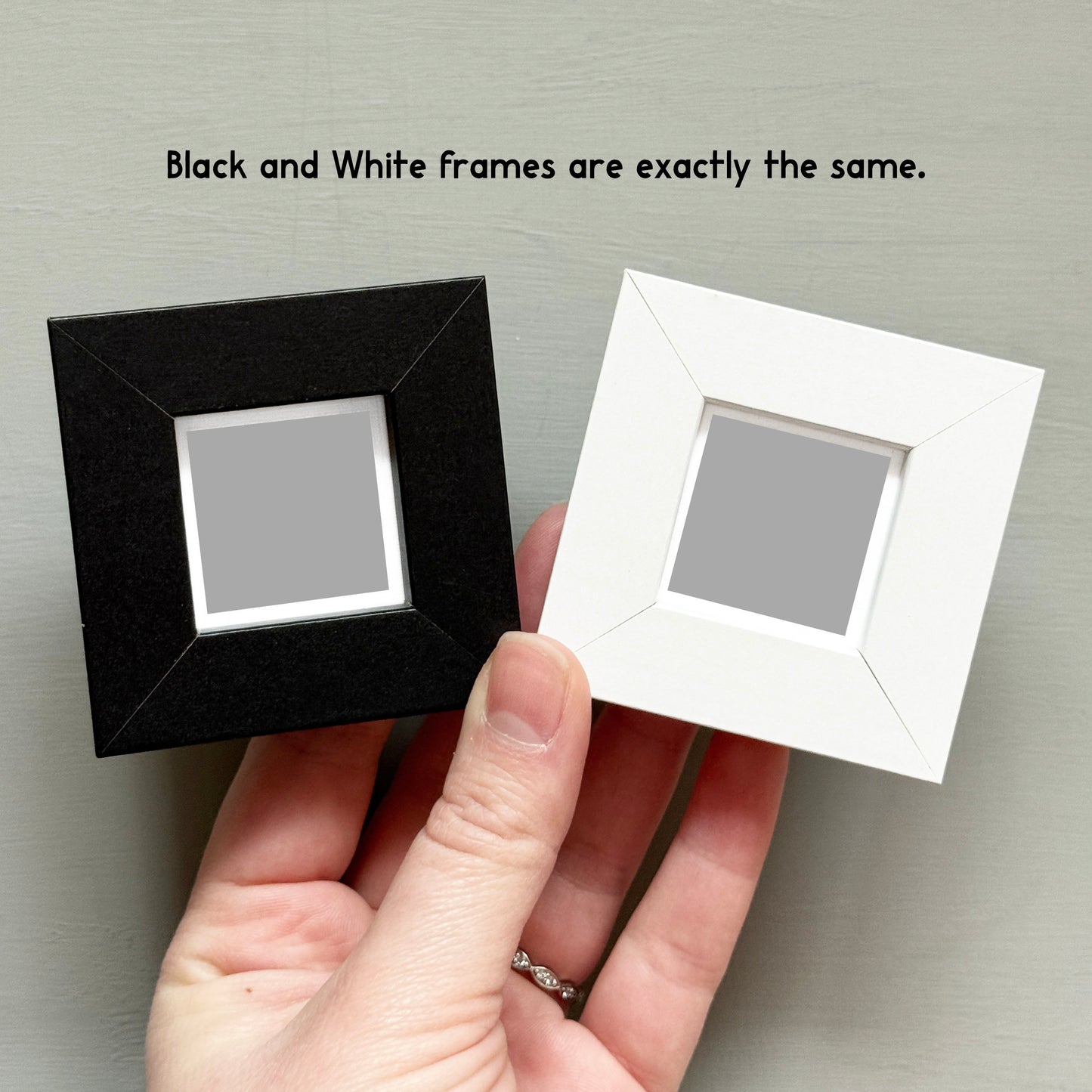 A hand holding two square picture frames, one black and one white, against a grey background.