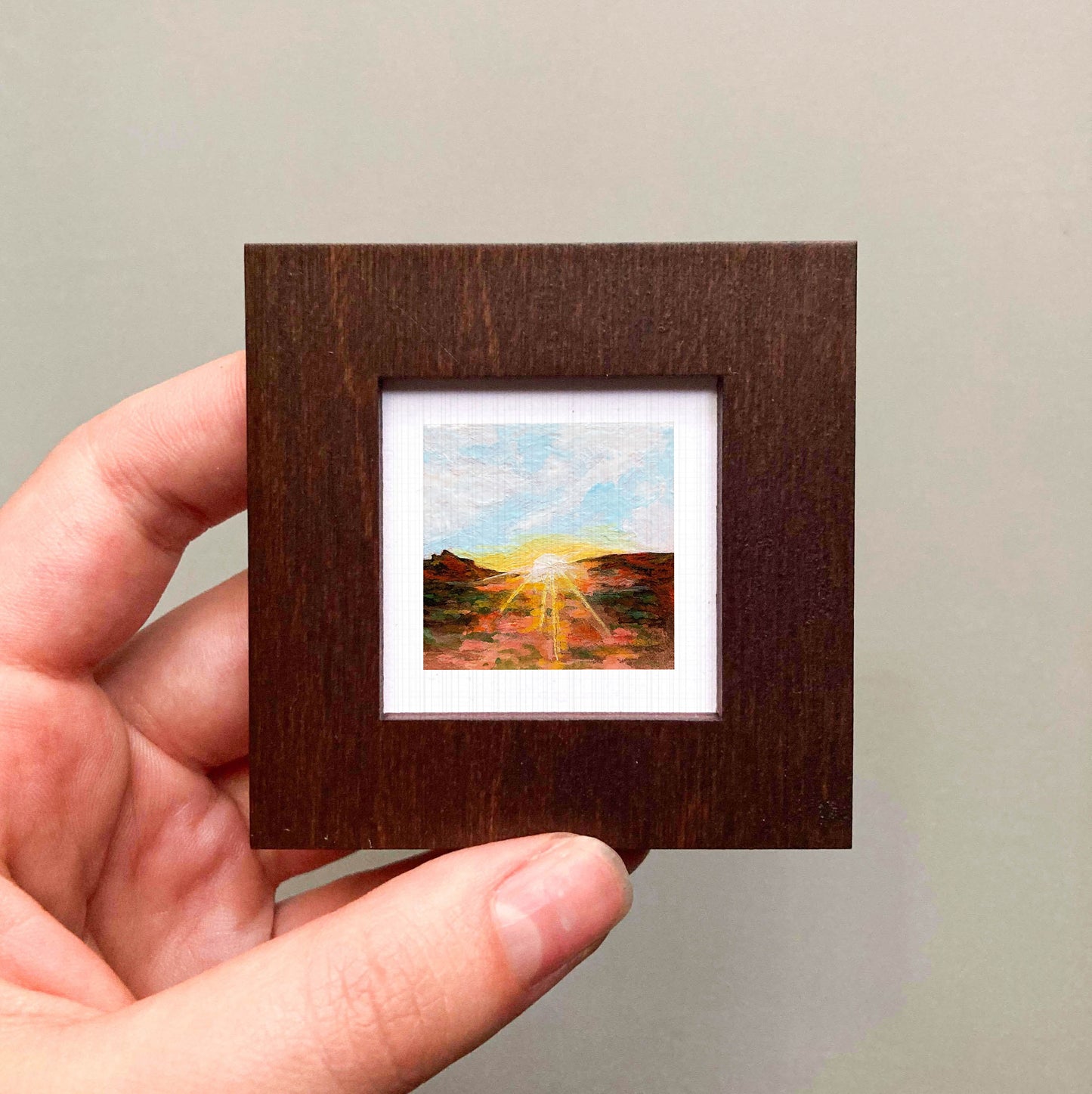 A hand is holding a small wooden frame containing a miniature painting of a landscape with a sunset.