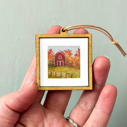 A hand holding a small wooden frame containing a painting of a red barn surrounded by autumn foliage.