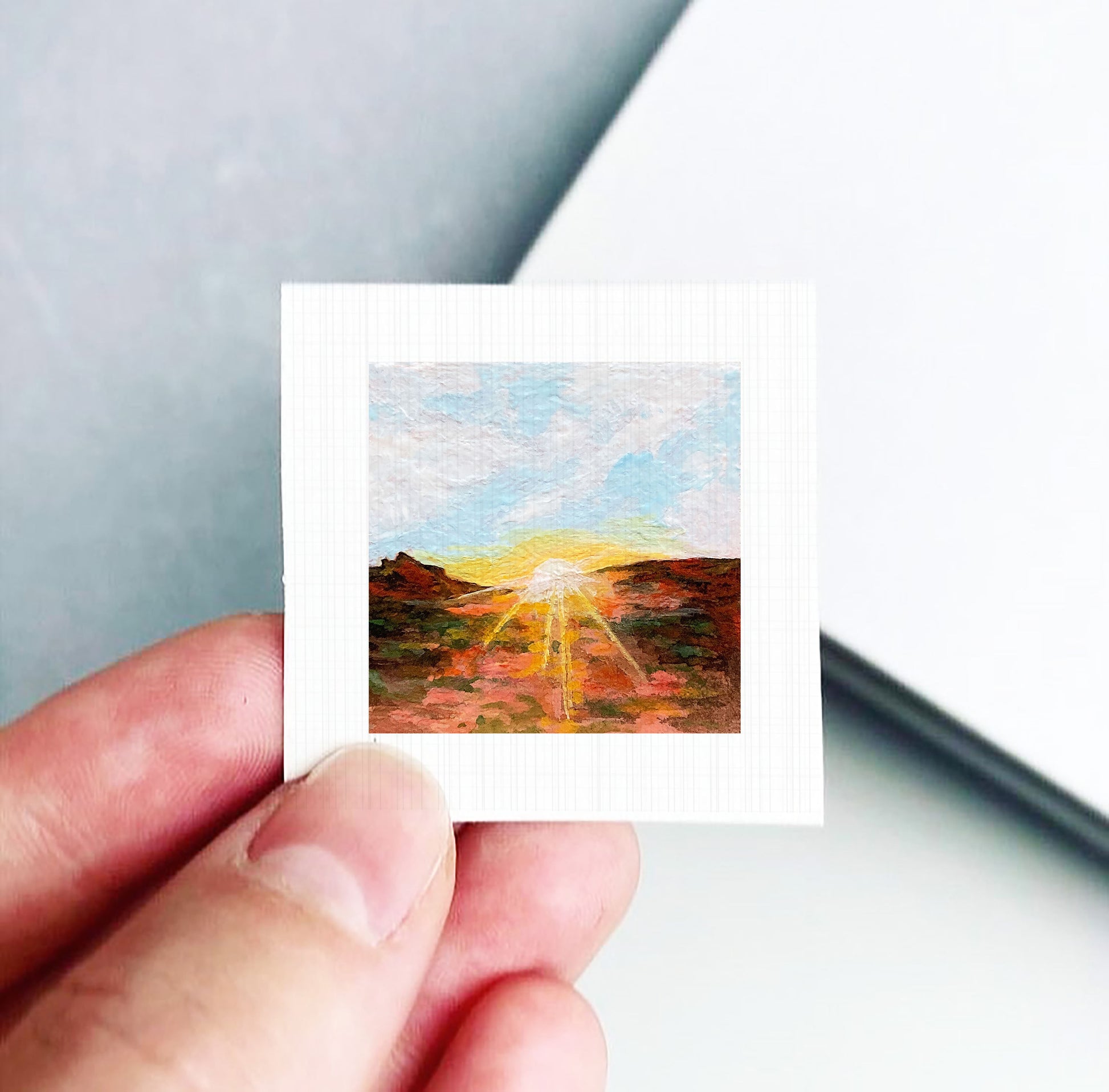 A hand holding a small square painting depicting a landscape with a sunset over a mountain.
