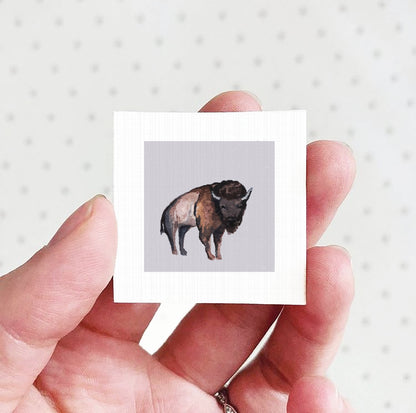 A hand holding a small square picture of a bison.