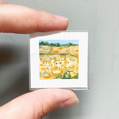 A person's hand holding a small square picture of a landscape with buildings and trees.