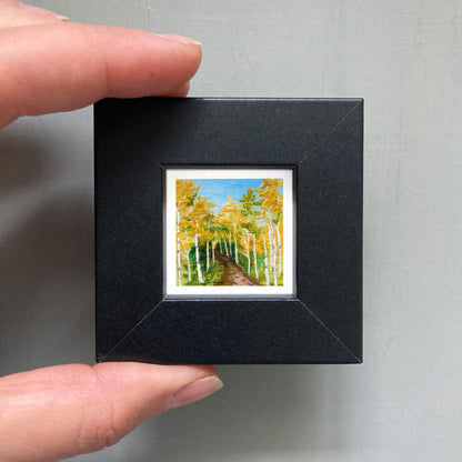 A person's hand holding a small black picture frame containing a painting of a forest with yellow trees.