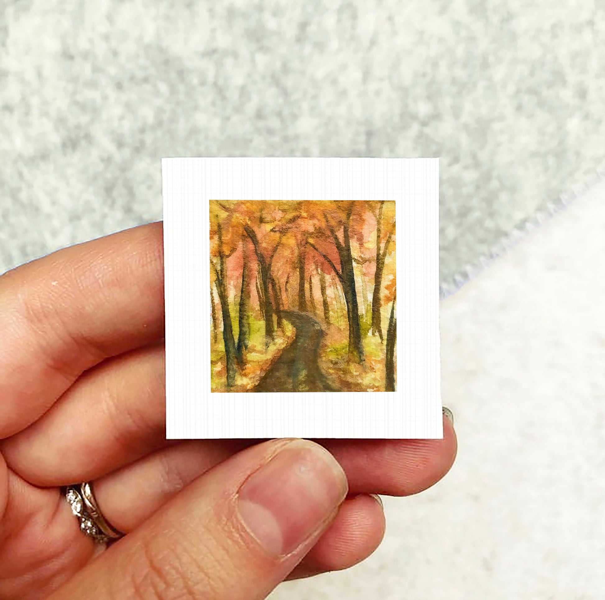 A hand holding a small square painting depicting a forest scene with a winding path through the trees.