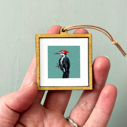 A person's hand holding a small wooden frame containing a framed image of a woodpecker.