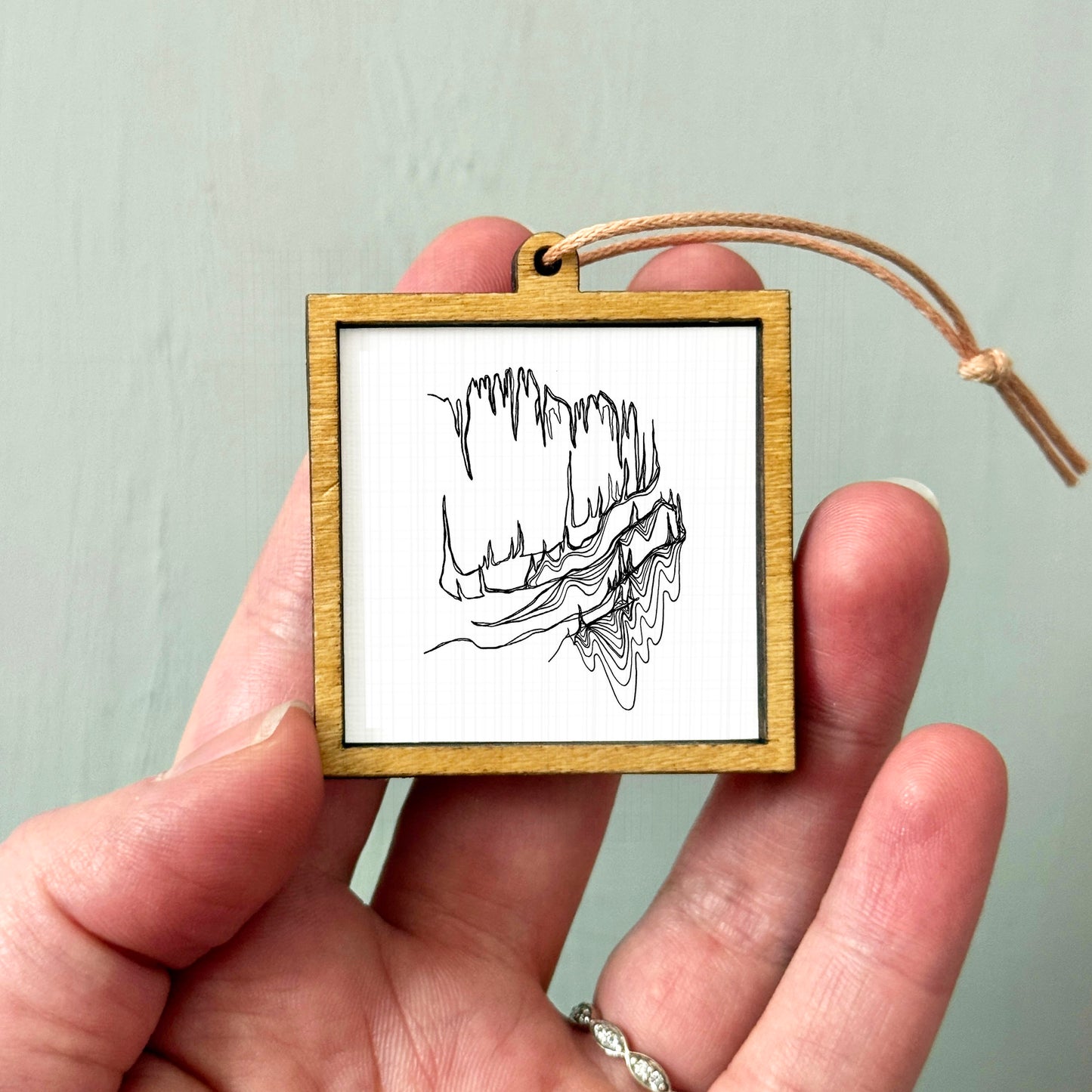 A hand holding a small wooden frame containing a black and white line drawing of a landscape.