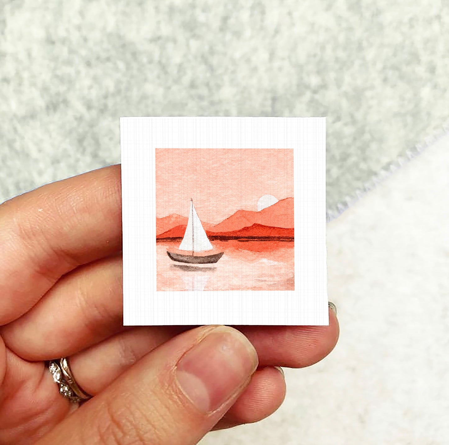 A hand holding a small square artwork depicting a sailboat on a body of water with mountains in the background.