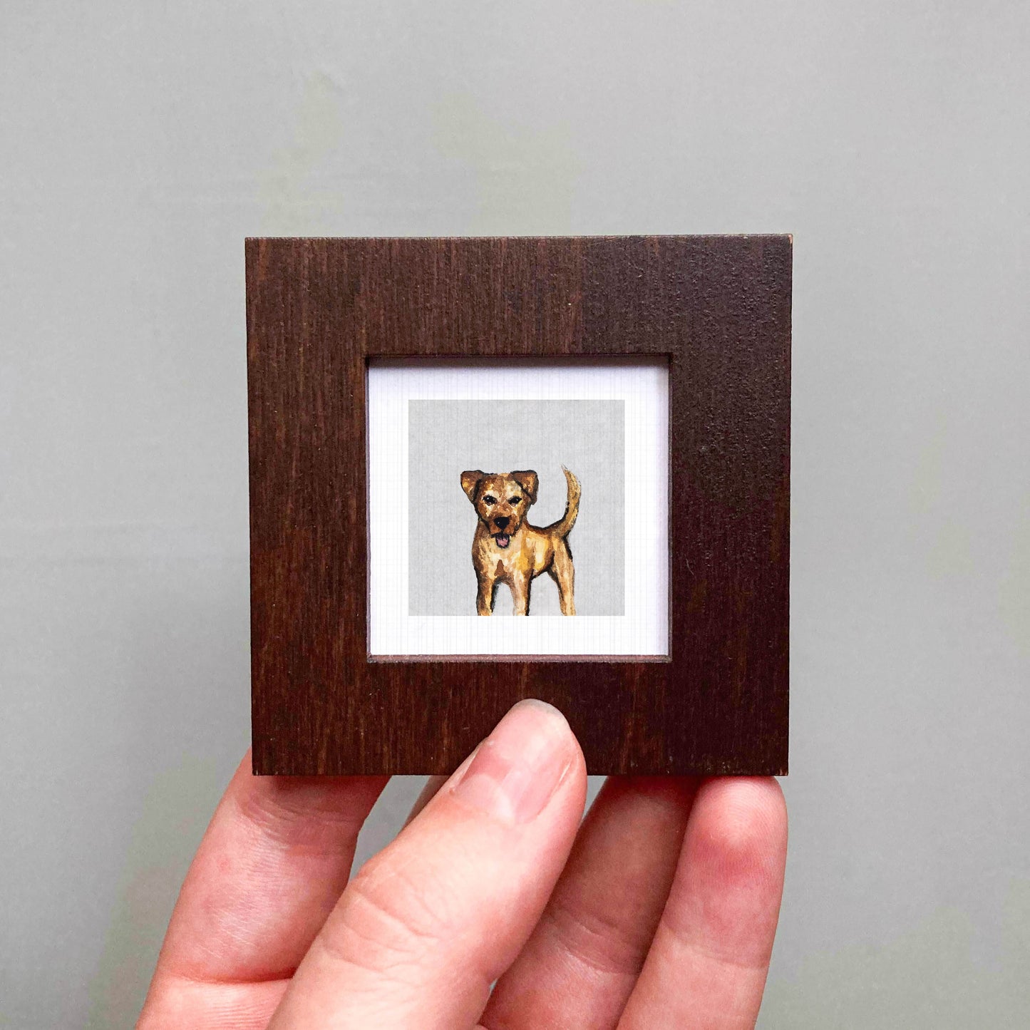 A person's hand holding a small wooden frame containing a painting of a dog.