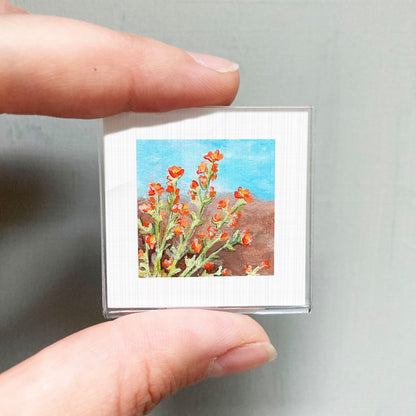 A person's hand holding a small square picture of a field of red flowers against a blue sky.