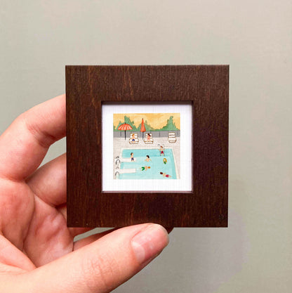 A hand is holding a small, square-shaped picture frame containing an illustration of a swimming pool with people enjoying the water.