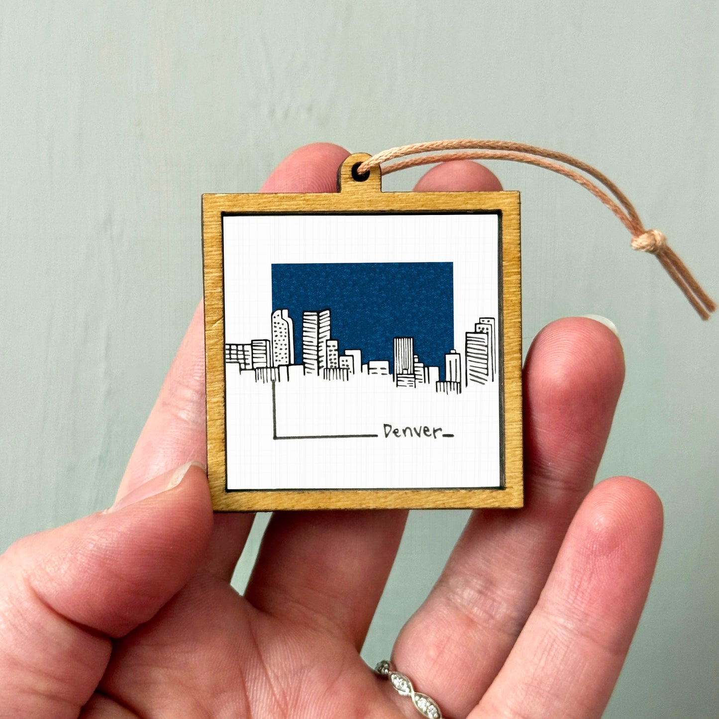 A hand holding a small wooden frame with a picture of a city skyline and the word " Denver" written on it.