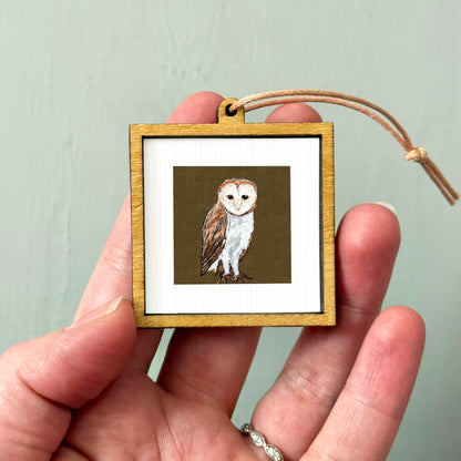 A person's hand holding a small wooden frame containing a picture of an owl.