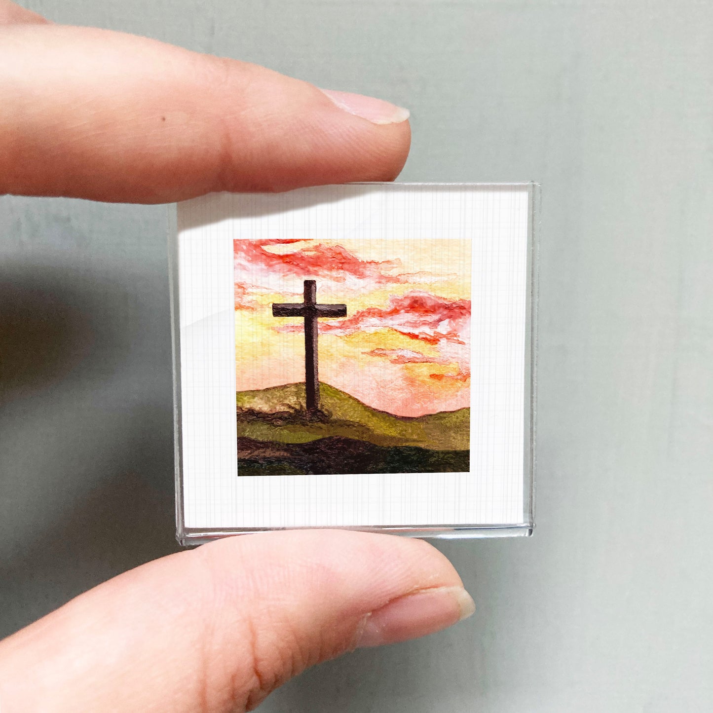 A person's hand is holding a small square-shaped object that displays a painting of a cross against a colorful sky.