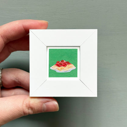 A hand is holding a small white picture frame containing an illustration of a plate of spaghetti with tomato sauce.