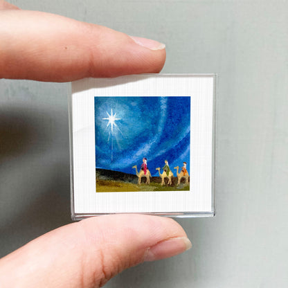 A hand holding a small square picture depicting a scene with three people and a star in the sky.