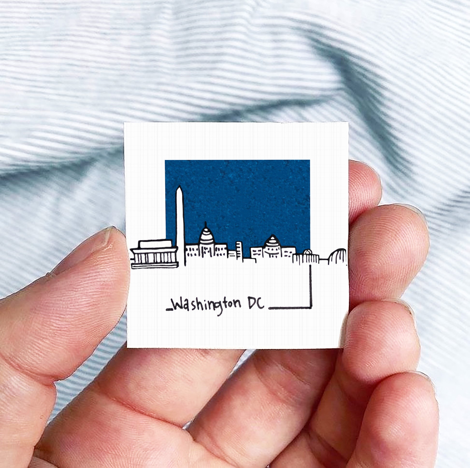 A hand holding a small square-shaped object with a drawing of Washington, D.C. on it.