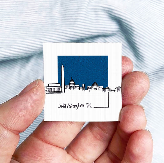 A hand holding a small square-shaped object with a drawing of Washington, D.C. on it.