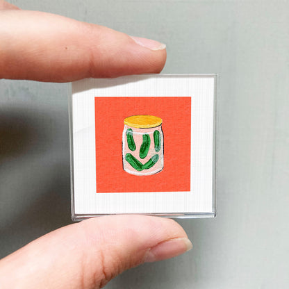 A person's hand holding a small square with an illustration of a jar of pickles on it against a red background.