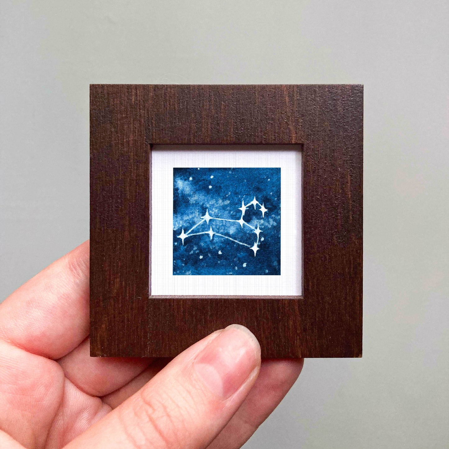 A hand is holding a small wooden frame containing a framed image of a starry night sky.