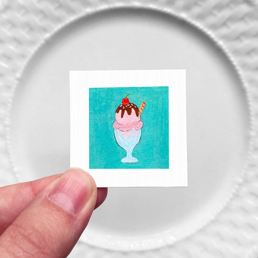 A hand is holding a small square picture of a colorful ice cream cone with a cherry on top.