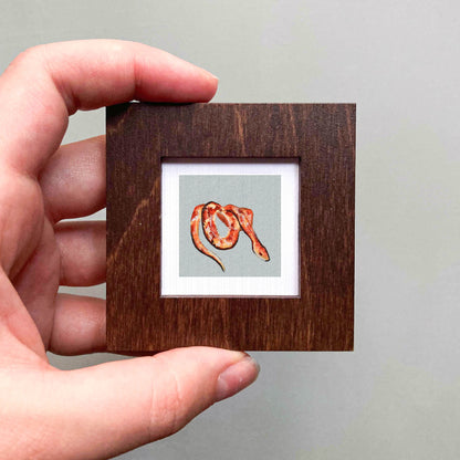 A hand is holding a small wooden frame containing a painting of a snake.
