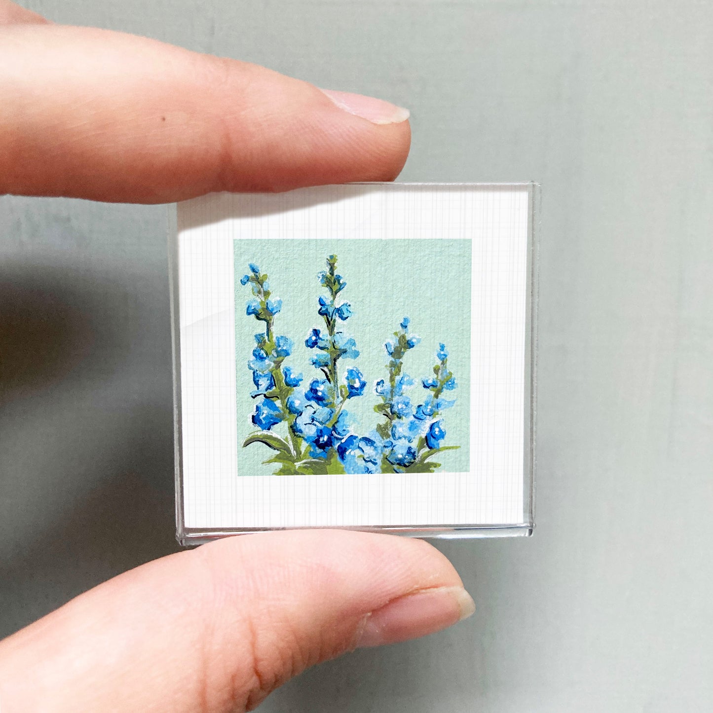 A person's hand holding a small square picture of blue flowers against a light green background.
