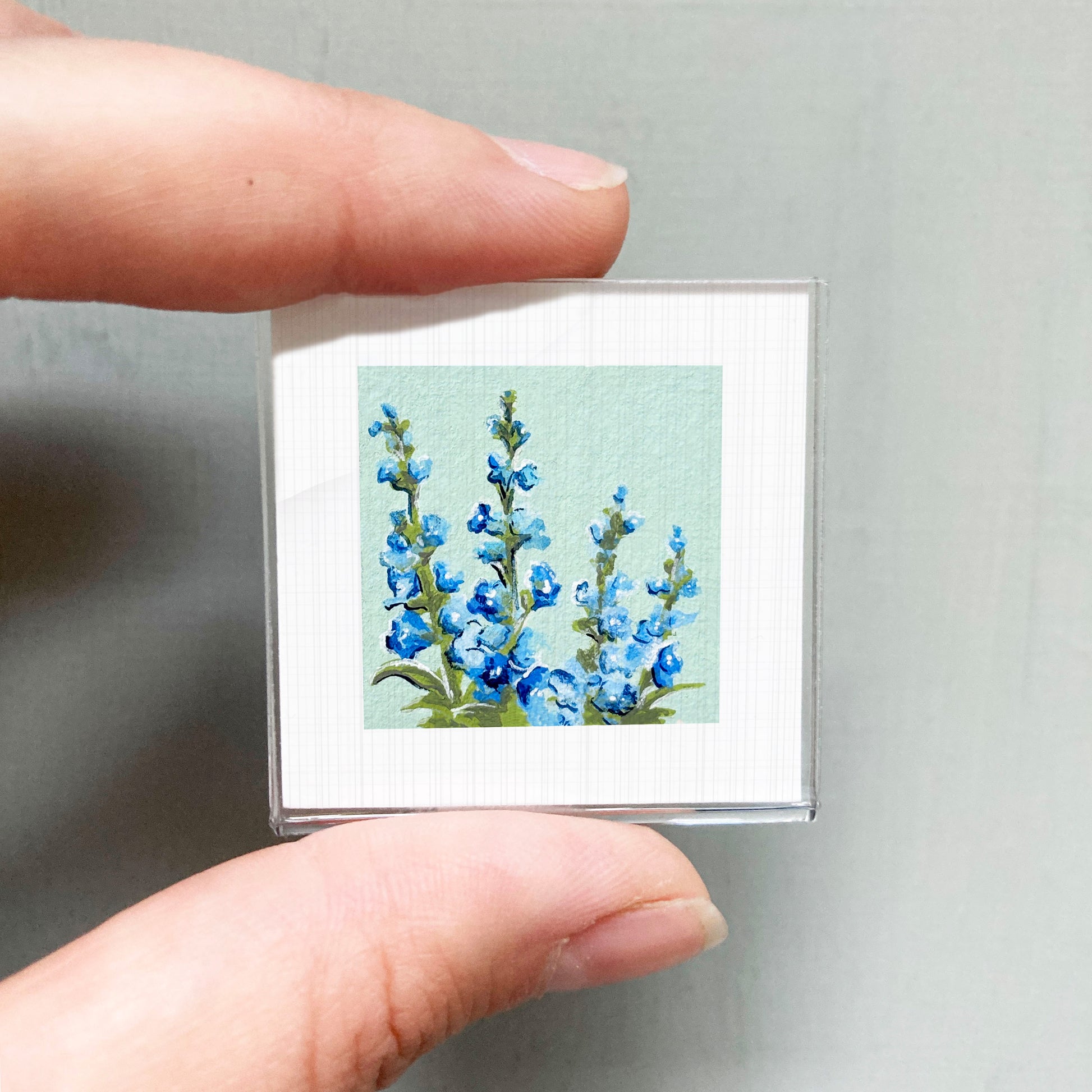 A person's hand holding a small square picture of blue flowers against a light green background.