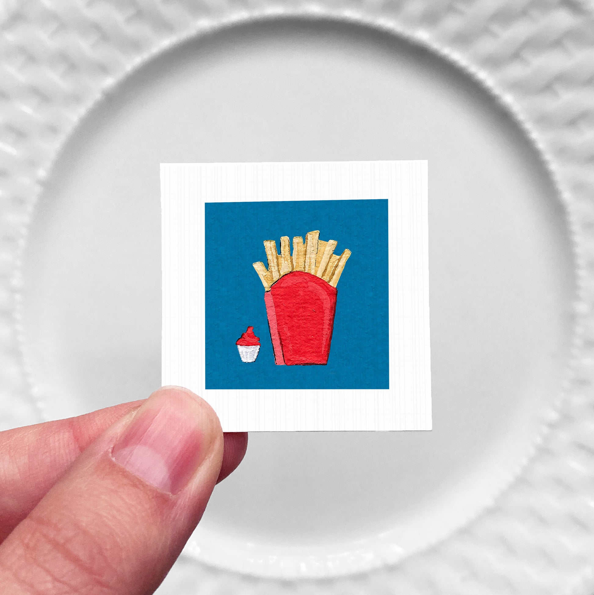 A hand is holding a small square with a red container of french fries and a cup of ketchup.
