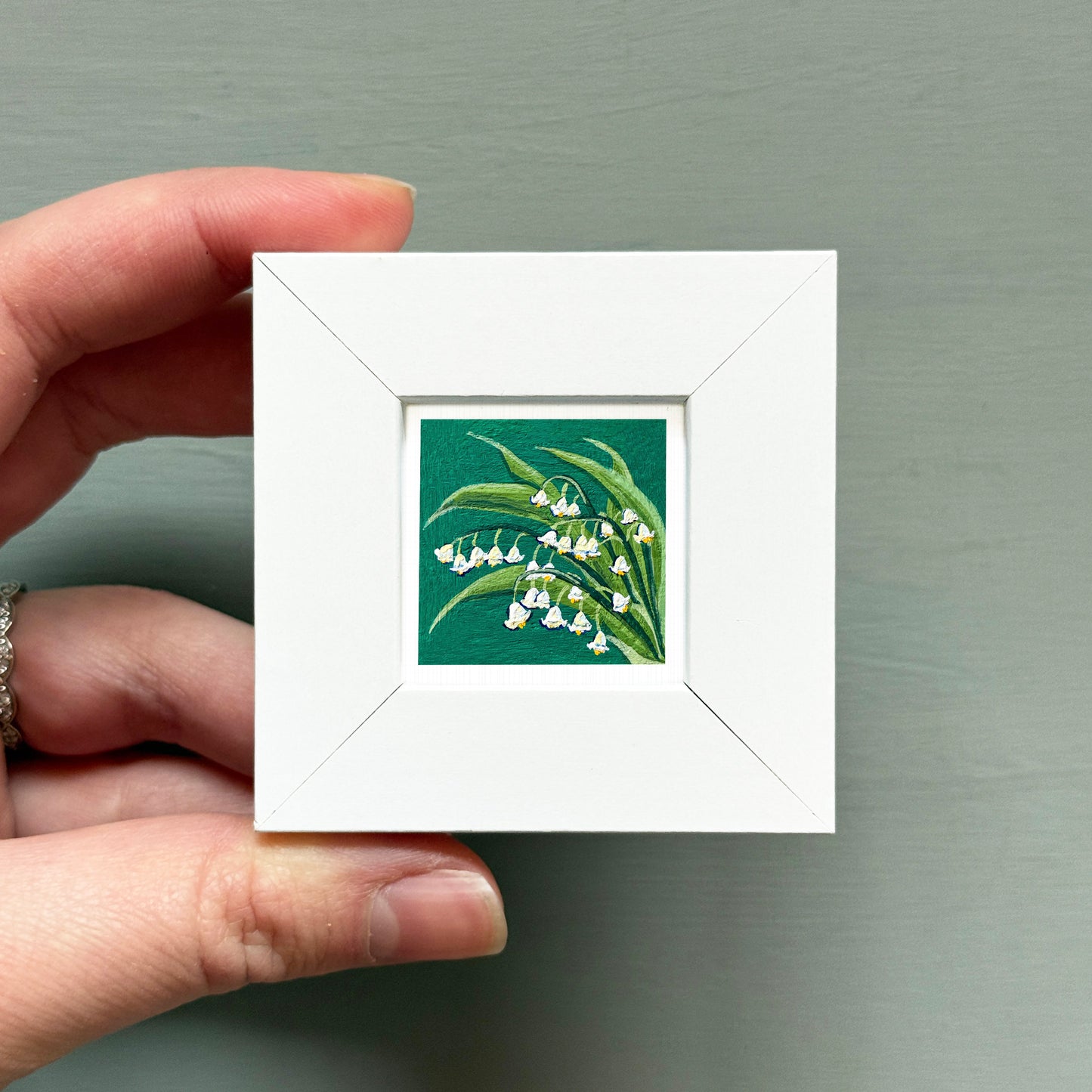 A hand is holding a small white square frame containing a painting of lily of the valley flowers.