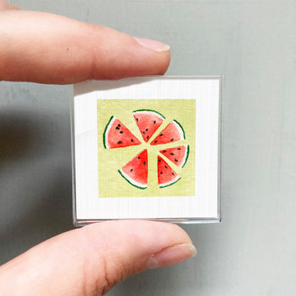 A hand is holding a small square with a watermelon slice design on it.