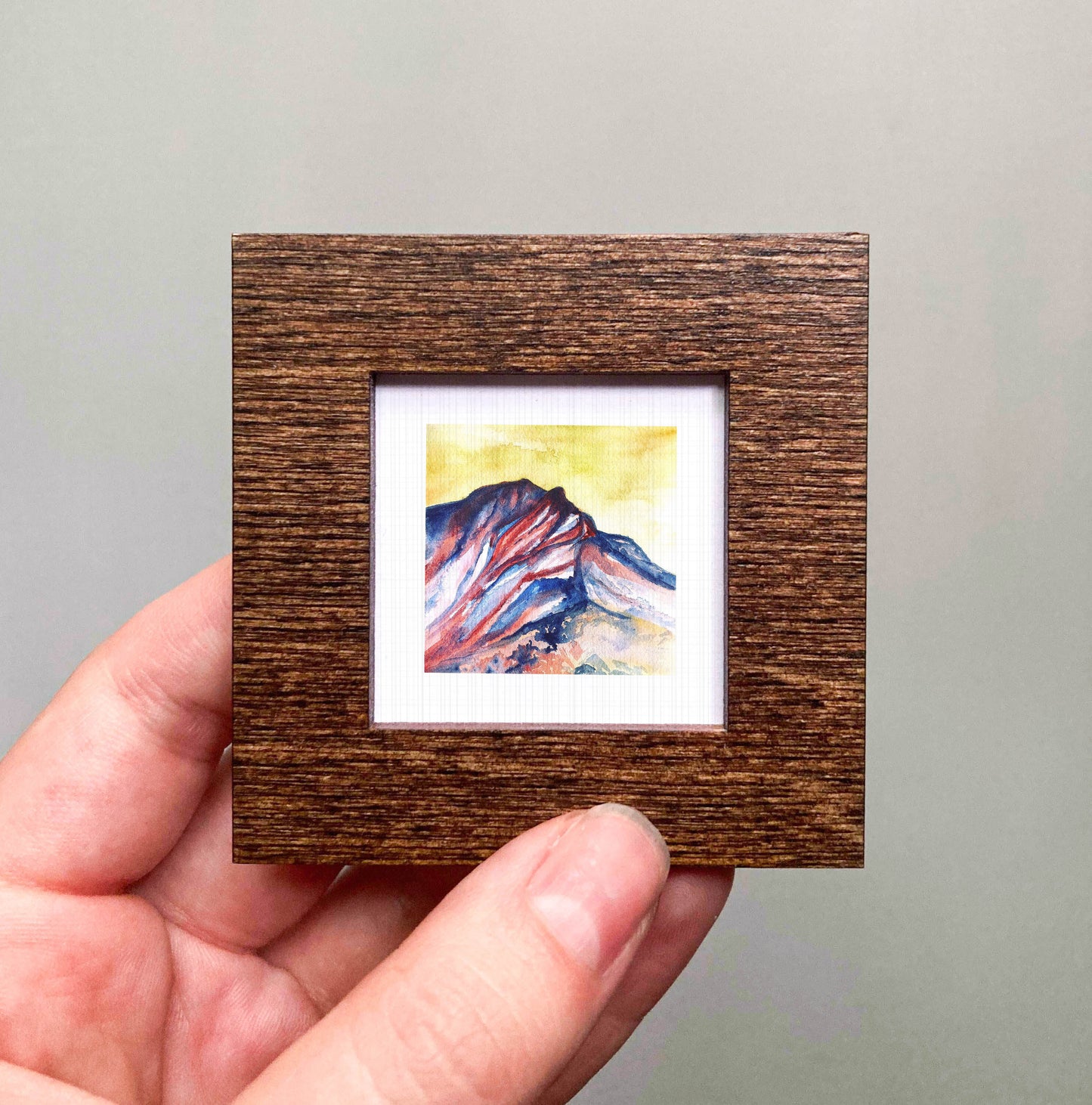 A hand is holding a small wooden frame containing a watercolor painting of a mountain landscape.
