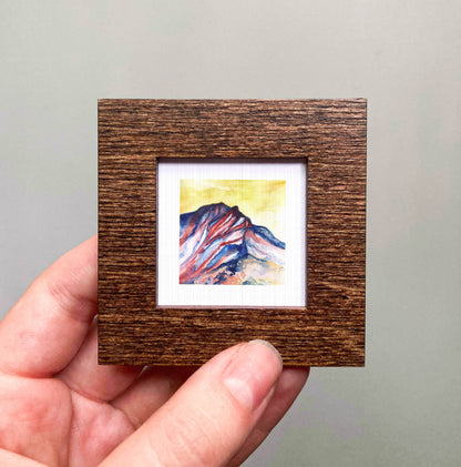 A hand is holding a small wooden frame containing a watercolor painting of a mountain landscape.