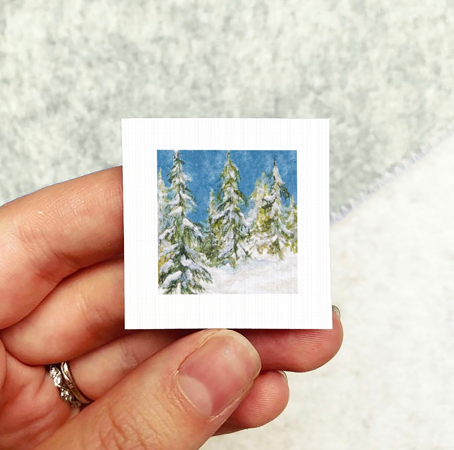 A hand holding a small square card with a painting of snow-covered pine trees on it.