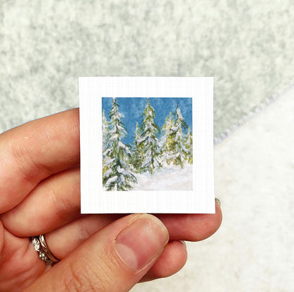 A hand holding a small square card with a painting of snow-covered pine trees on it.