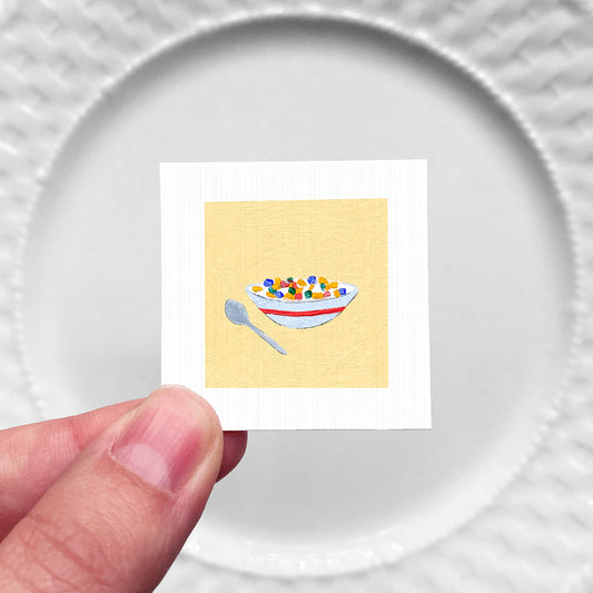A hand is holding a small square with a picture of a bowl of cereal on it, against a white background.