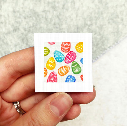 A hand holding a small square card with a colorful pattern of Easter eggs on it.