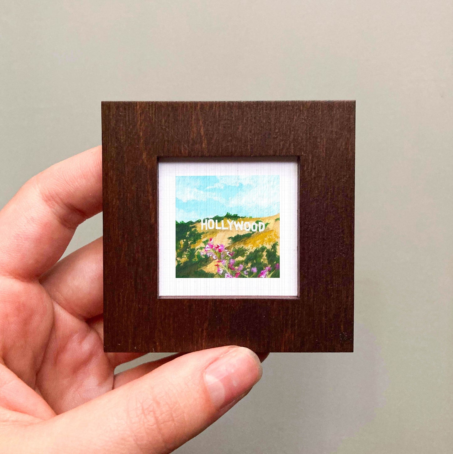 A hand is holding a small, square-shaped picture frame containing a painting of Hollywood.